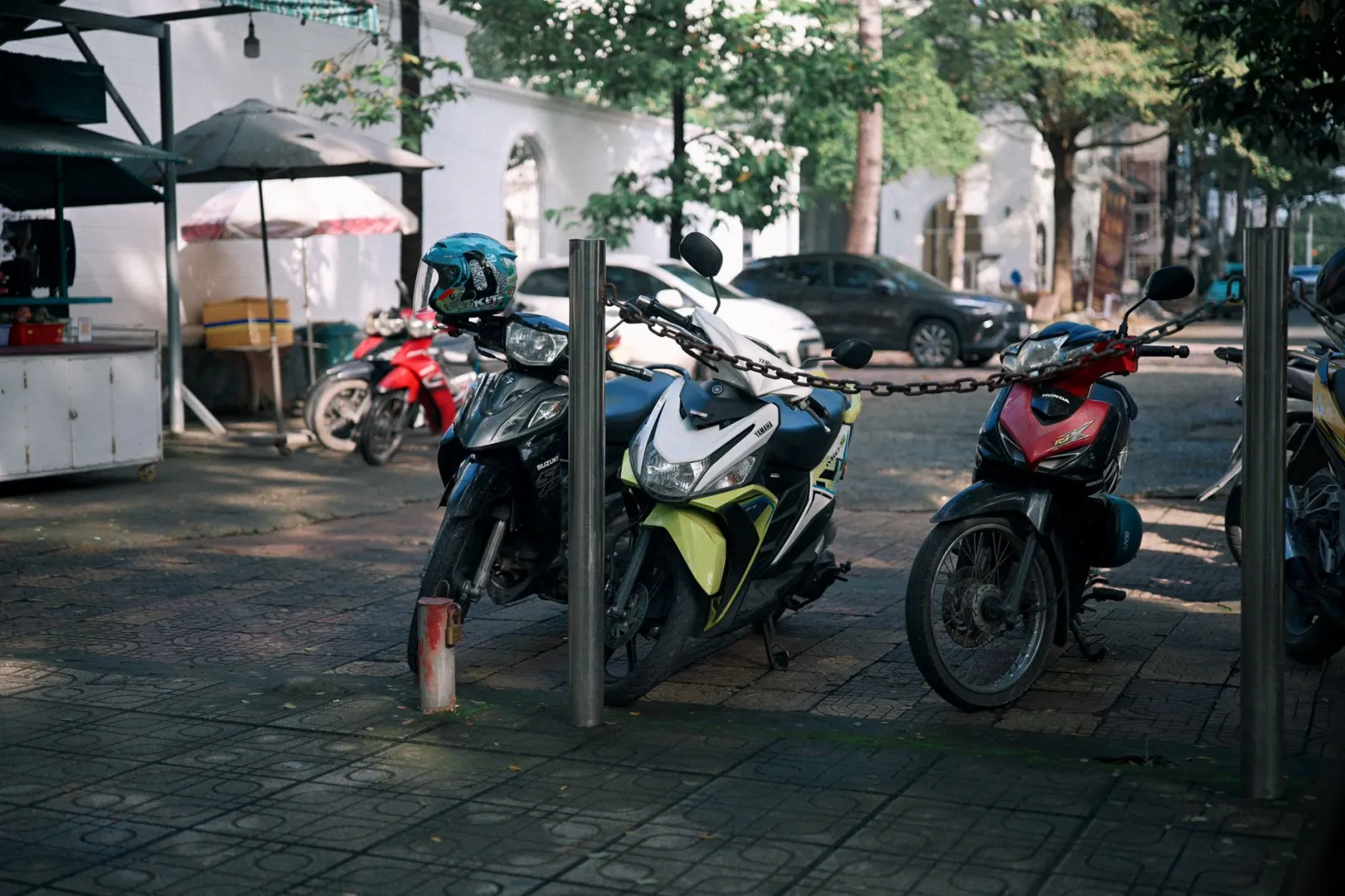 Three motorcycles chained together on a city sidewalk, shaded by trees and near a food stall.